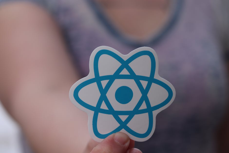 A close-up of a person holding a sticker with a blue atom symbol, with the person's hand and a blurred background showing a person's torso wearing a purple top. The focus is on the atom sticker, which